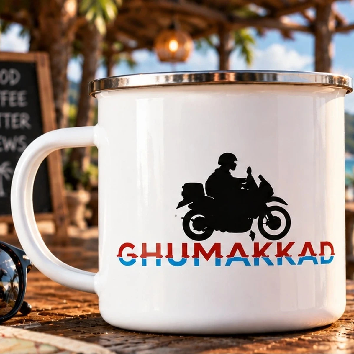 GHUMAKKAD 300ml enamel camping mug with biker silhouette and red-blue lettering on a beachside cafe table