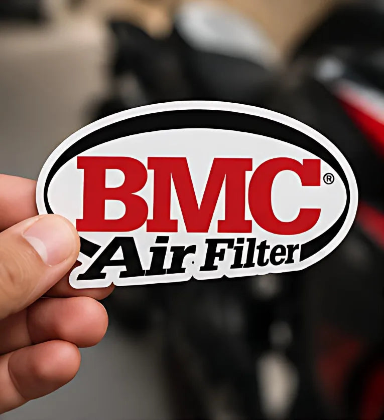 Hand holding BMC Air Filter 4 inch sticker in front of a motorcycle